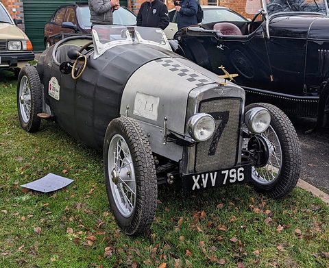 A grey and silver boat-tailed two-seat special with wire wheels on grass among parked cars. Opens a larger view