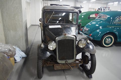 Head-on view of a small dark saloon with a narrow radiator in a museum, blue cars behind. Opens a larger view