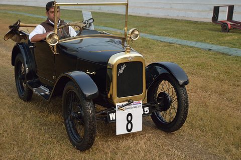 A small dark open tourer with a brass radiator and rally number 8, its driver at the wheel on a grass field. Opens a larger view