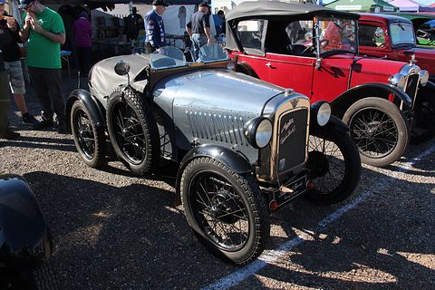 A silver-grey sports two-seater with a pointed tail and cycle-type wings in a paddock with spectators. Opens a larger view