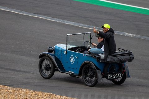 A racing driver in a cap waves from the back of a small blue open tourer on a race track. Opens a larger view