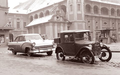 A sepia winter street: a tall upright small coupé with wire wheels beside a two-tone 1950s saloon before baroque buildings. Opens a larger view