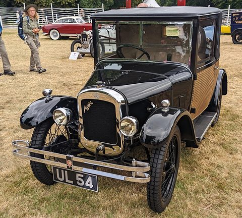 A black and varnished-wood upright saloon with a brass radiator and a front bumper, on grass at a show. Opens a larger view