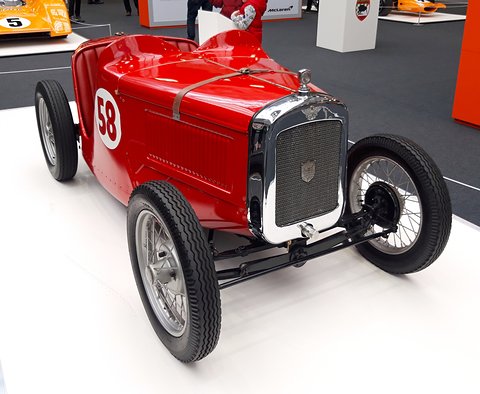 A red pointed-tail racing two-seater with the number 58 and wire wheels on a white show floor. Opens a larger view