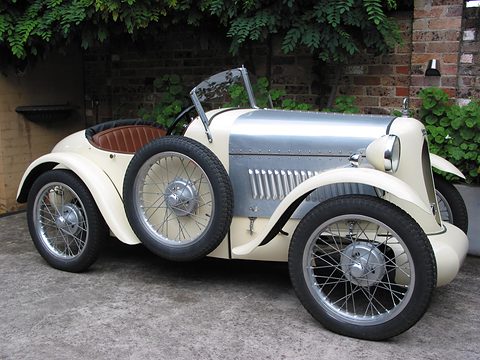 A cream and silver two-seater with flowing wings, louvred bonnet and wire wheels before a brick wall. Opens a larger view