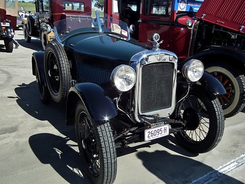 A dark green speedster with a chrome radiator and wire wheels in a sunny paddock beside red cars. Opens a larger view
