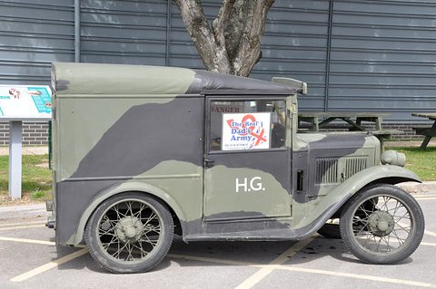 A camouflaged grey-green van with H.G. on its side parked outside a corrugated shed. Opens a larger view