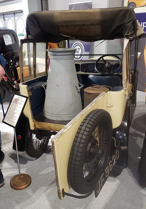 A cream open delivery car with a tall milk churn in the back and its door open, on a show stand. Opens a larger view