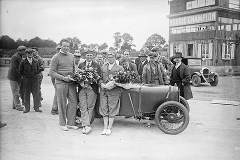 Black and white photograph of three people holding bouquets beside a small open racing car, a grandstand behind. Opens a larger view