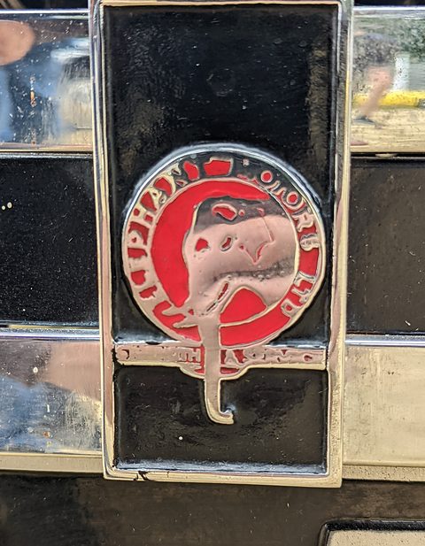 A round red enamel badge with a white emblem mounted on a black and gold radiator shell. Opens a larger view