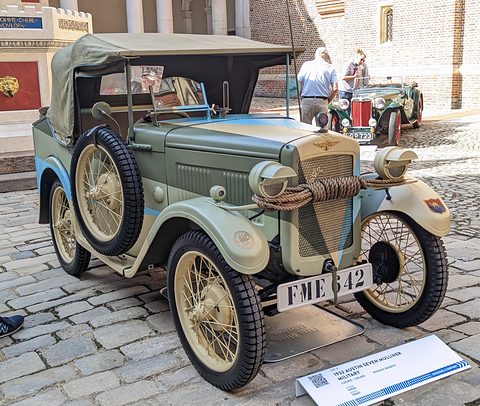 A pale green open utility car with a spare wheel at the front, cream wheels and a black hood, on cobbles. Opens a larger view
