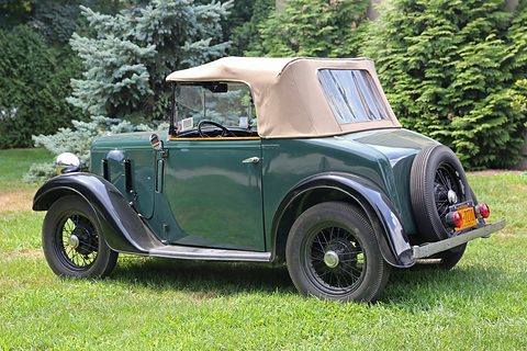 The rear quarter of a dark green two-seater with its beige hood up and a spare wheel, on grass. Opens a larger view
