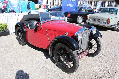 A red sports two-seater with black wings and a cowled radiator in a gravel courtyard. Opens a larger view
