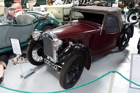 A maroon two-seater with its hood up and wire wheels behind a rope in a museum. Opens a larger view