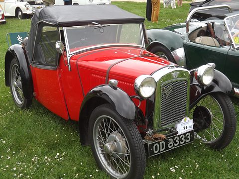 A red sports two-seater with a black hood up, a cowled radiator and wire wheels on grass. Opens a larger view