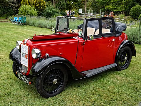 A red open two-seater with black wings and wheels on a lawn by flower beds. Opens a larger view