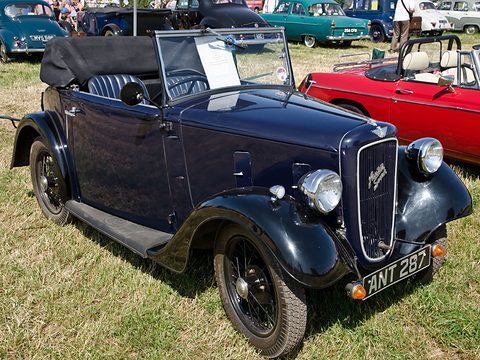 A dark blue two-seat tourer with its hood folded and a cowled grille on grass among cars. Opens a larger view