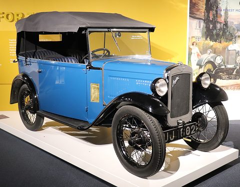 A blue four-seat tourer with its black hood up and wire wheels on a white plinth before a yellow wall. Opens a larger view