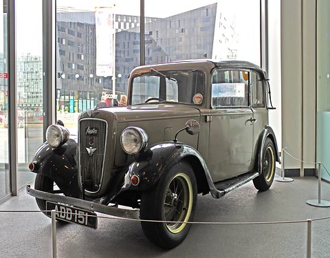 A dark cabriolet with its hood up and spoked wheels inside a glass-walled foyer. Opens a larger view