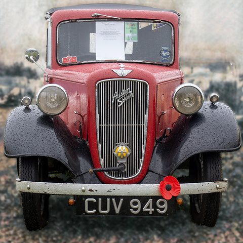 Head-on view of a red car with a cowled grille, chrome headlamps and a poppy on its bumper. Opens a larger view