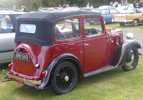The rear quarter of a red cabriolet with a black hood up and black wheels, parked on grass. Opens a larger view