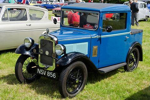 A small blue pick-up with a cab, black wings and black wheels on grass among other vehicles. Opens a larger view