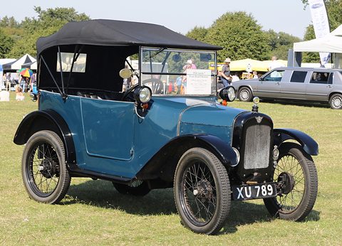 A blue open tourer with a tall black folding hood, black wings and wire wheels on grass at a show. Opens a larger view