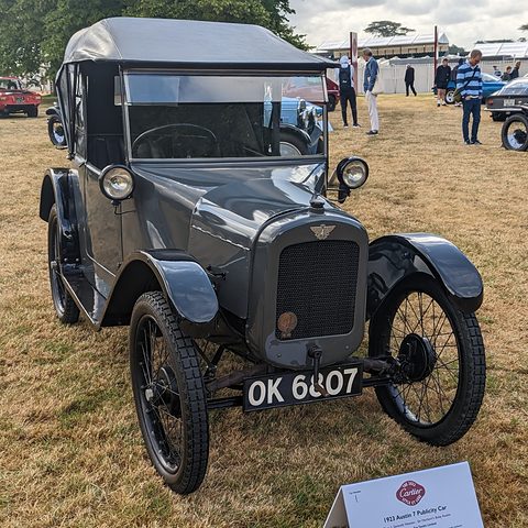A small dark grey open tourer with its hood up and wire wheels on grass, a display card in front. Opens a larger view