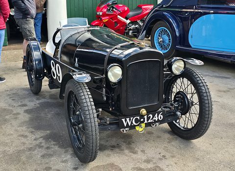 A narrow black open racing car with cycle wings, wire wheels and the number 99 on concrete among visitors. Opens a larger view