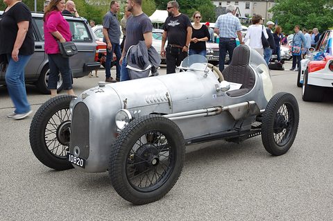 A grey single-seat racer with wire wheels in a car park, visitors around it. Opens a larger view