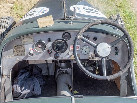 A worn aluminium dashboard with dials behind a large steering wheel in a green open racer. Opens a larger view