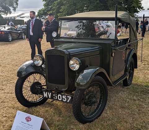 A small dark green tourer with a raised hood frame and aerials on top, men in suits behind it on grass. Opens a larger view