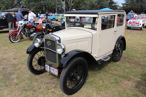 A cream square saloon with black wings and wheels on a grass field, motorcycles and people beyond. Opens a larger view
