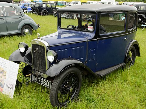 A dark blue square saloon with black wings and wire wheels on grass beside a sign board. Opens a larger view