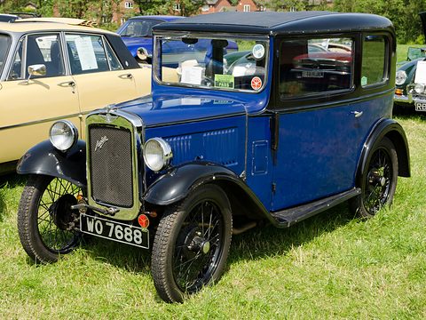 A blue and black square saloon with black wheels and a notice on its window, on grass. Opens a larger view