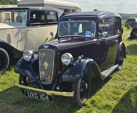 A dark rounded saloon with a cowled chrome grille and wire wheels on grass beside a cream car. Opens a larger view