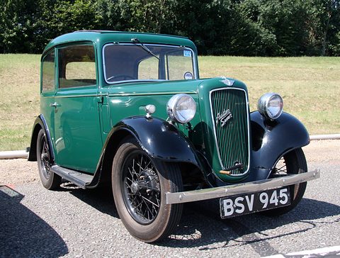 A green rounded saloon with a cowled grille and wire wheels on tarmac before a hedge. Opens a larger view