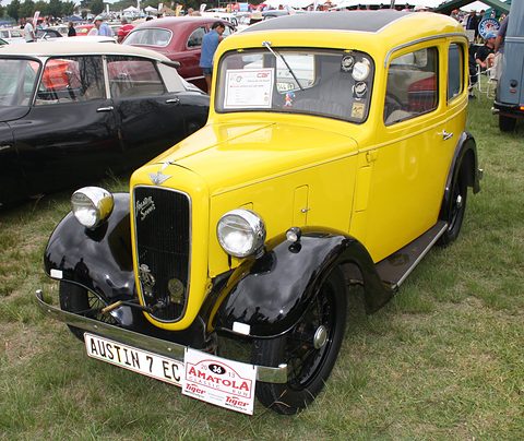 A yellow rounded saloon with black wings and a cowled grille on grass among other cars. Opens a larger view