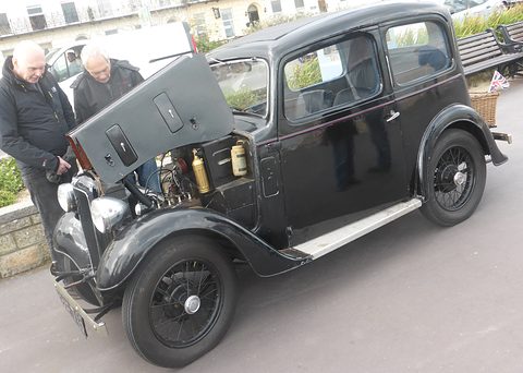 A black rounded saloon with one side of its bonnet raised, two men looking at the engine. Opens a larger view