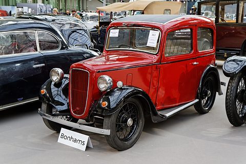 A red rounded saloon with black wings and wheels behind a Bonhams sign among other cars. Opens a larger view