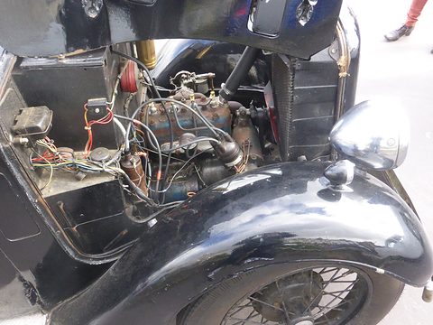 Close view of a small four-cylinder engine with wiring and a carburettor under a raised black bonnet. Opens a larger view