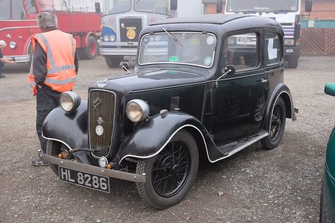 A black rounded saloon with a cowled grille on a gravel yard, a steward in a high-visibility vest behind. Opens a larger view