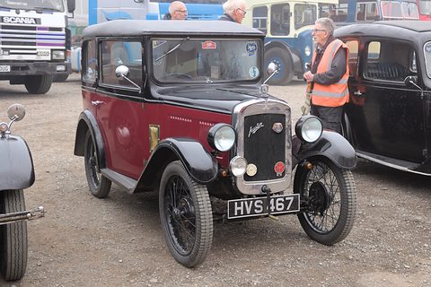 A maroon saloon with black wings and a chrome radiator shell on gravel, a man in a high-visibility vest beside it. Opens a larger view