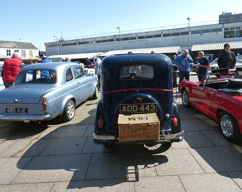 The rounded rear of a dark saloon with a wicker hamper strapped to the luggage grid, people and cars around. Opens a larger view