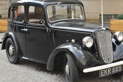 A black rounded saloon with a cowled grille and wire wheels on paving in a city square. Opens a larger view
