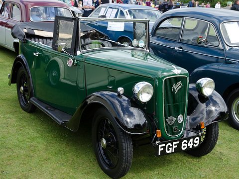 A dark green four-seat tourer with its hood folded and a cowled grille on grass among cars. Opens a larger view