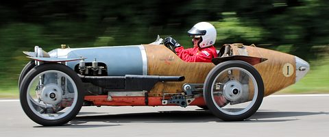 A long narrow special with a polished bonnet and a timber tail driven fast on a road course. Opens a larger view