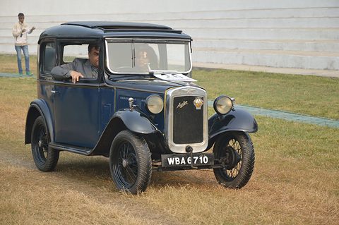 A dark blue square saloon with black wings and wire wheels on a dry grass field, its driver inside. Opens a larger view
