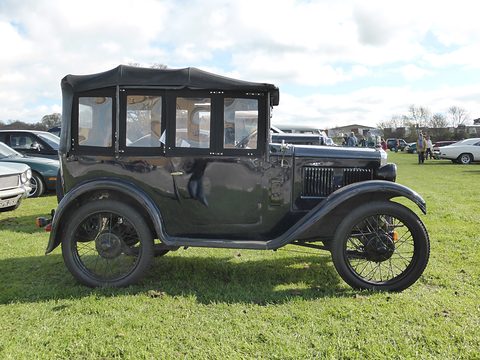 Side view of a black upright saloon with black wheels parked on grass, a cloudy sky behind. Opens a larger view