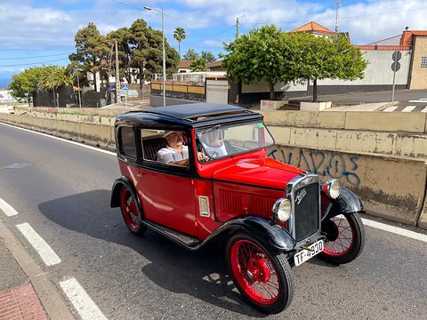 A small red saloon with a black roof driving on a sunny road lined with palms and a low wall. Opens a larger view
